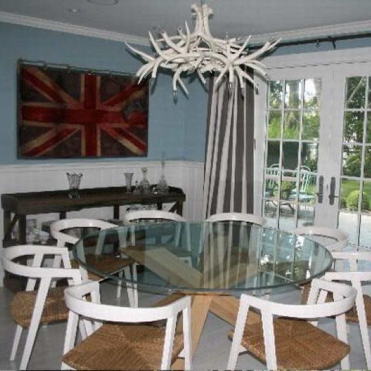 A dining area features a round glass table surrounded by white chairs with woven seats, alongside a British flag artwork and large glass doors leading to a garden.