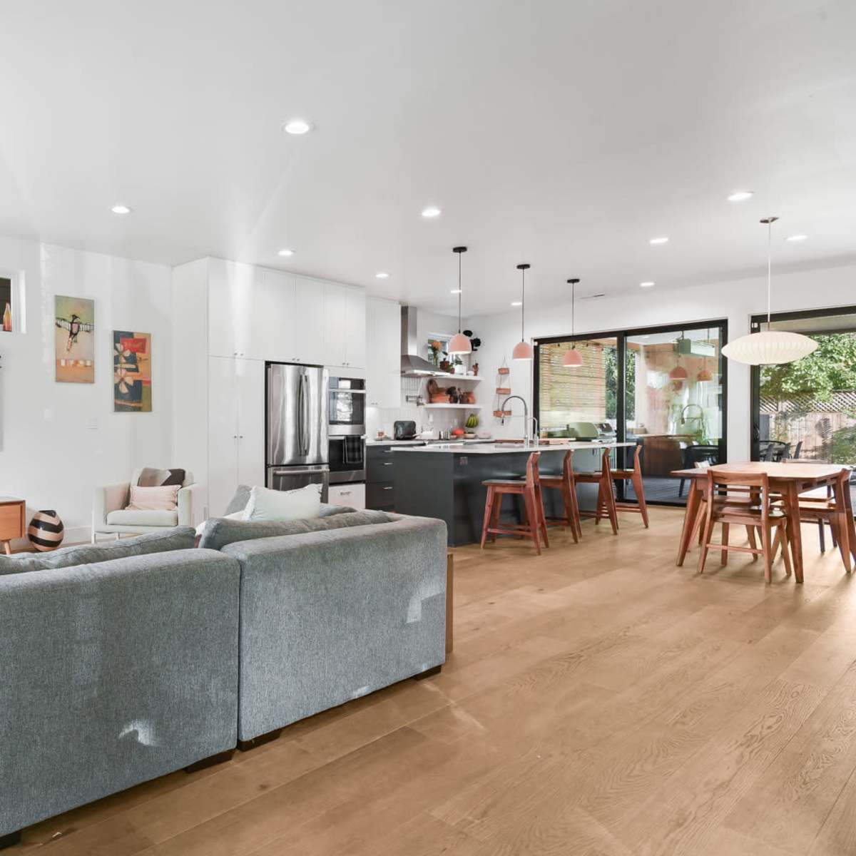 A modern living space features a gray sectional sofa, a dining area with a wooden table, and an open kitchen with black cabinetry and stainless steel appliances.