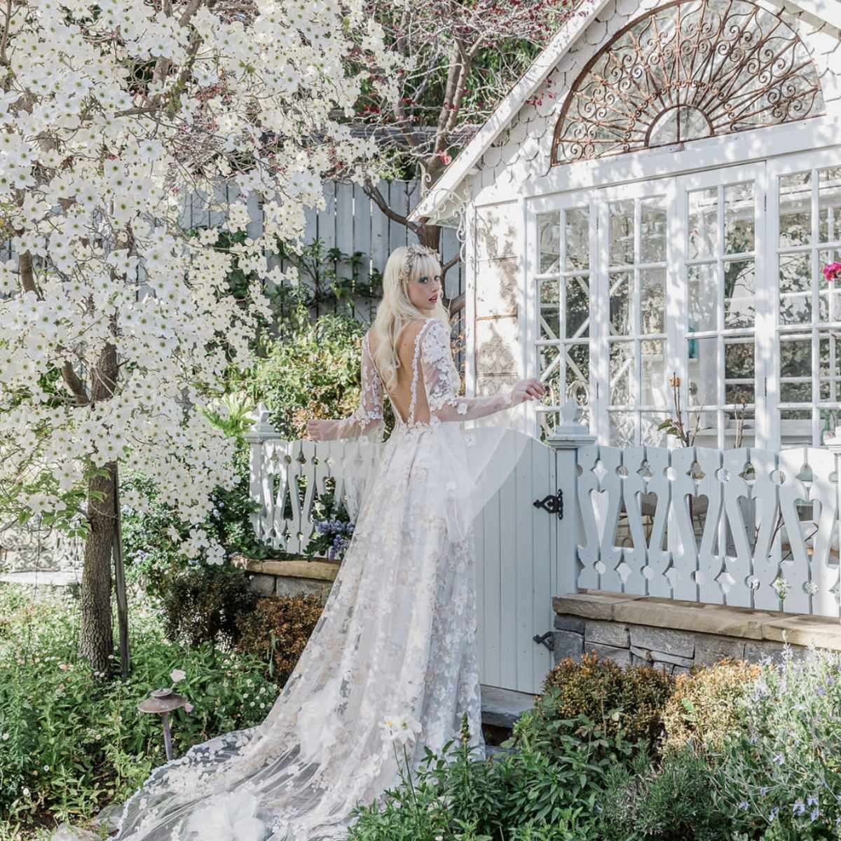 A woman in a detailed lace wedding dress stands by a vintage-style garden shed, surrounded by blooming white flowers and greenery.
