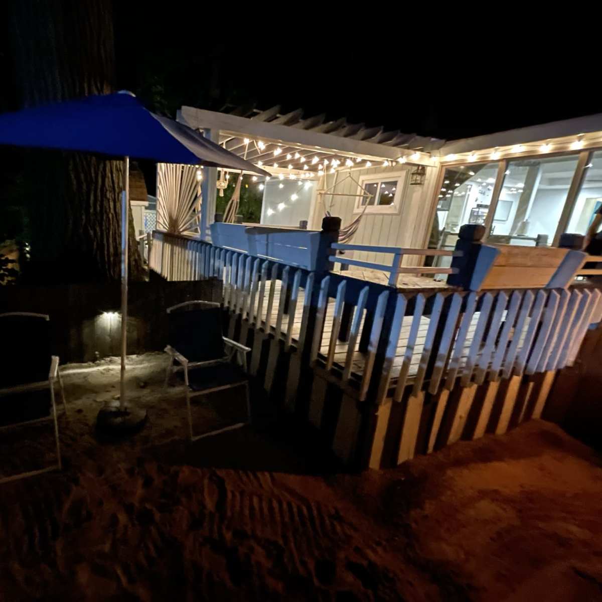 A sandy area beneath a blue umbrella with two chairs, adjacent to a well-lit deck and a house at night.