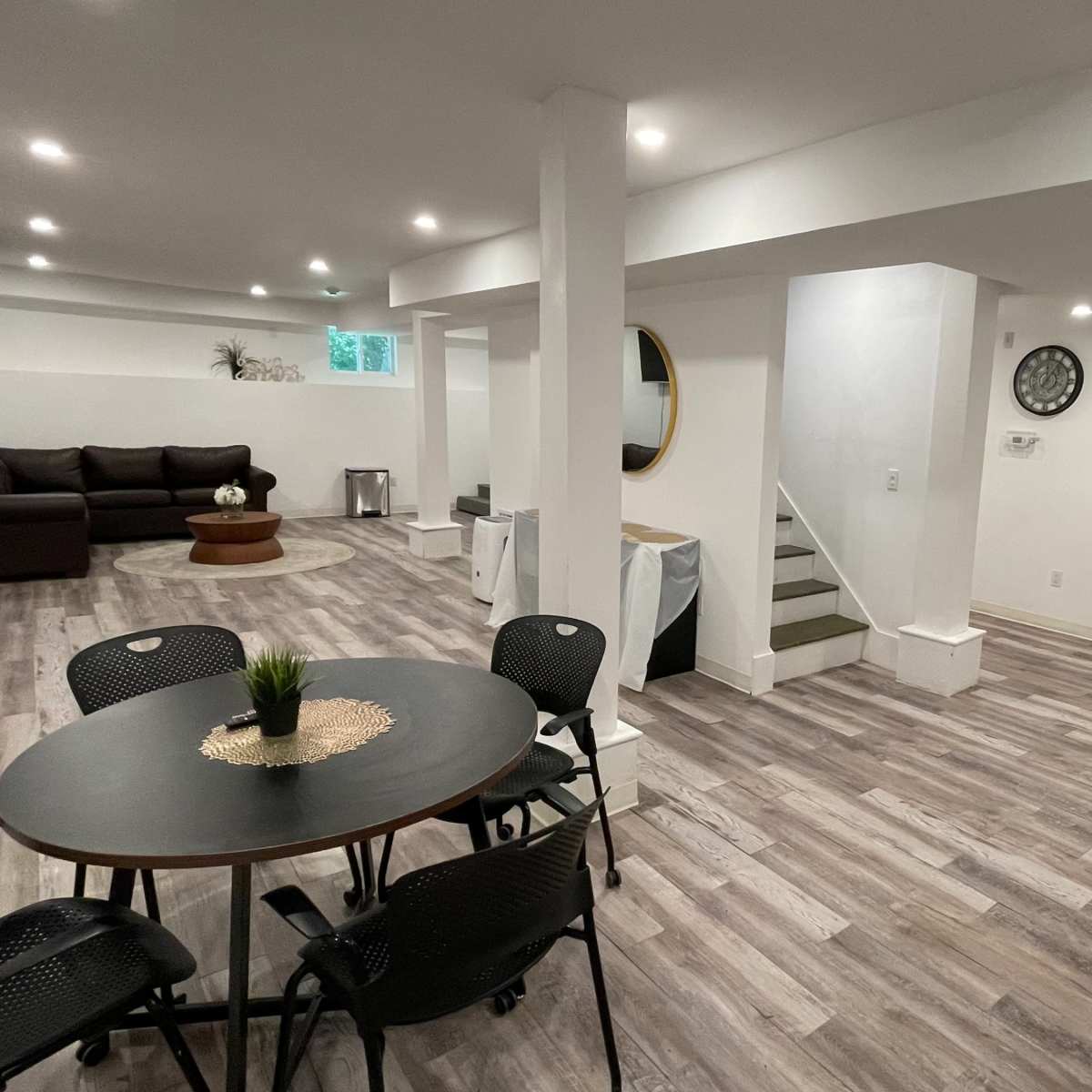 A modern, open-concept basement living space featuring a seating area, a dining table, and light-colored wooden flooring.