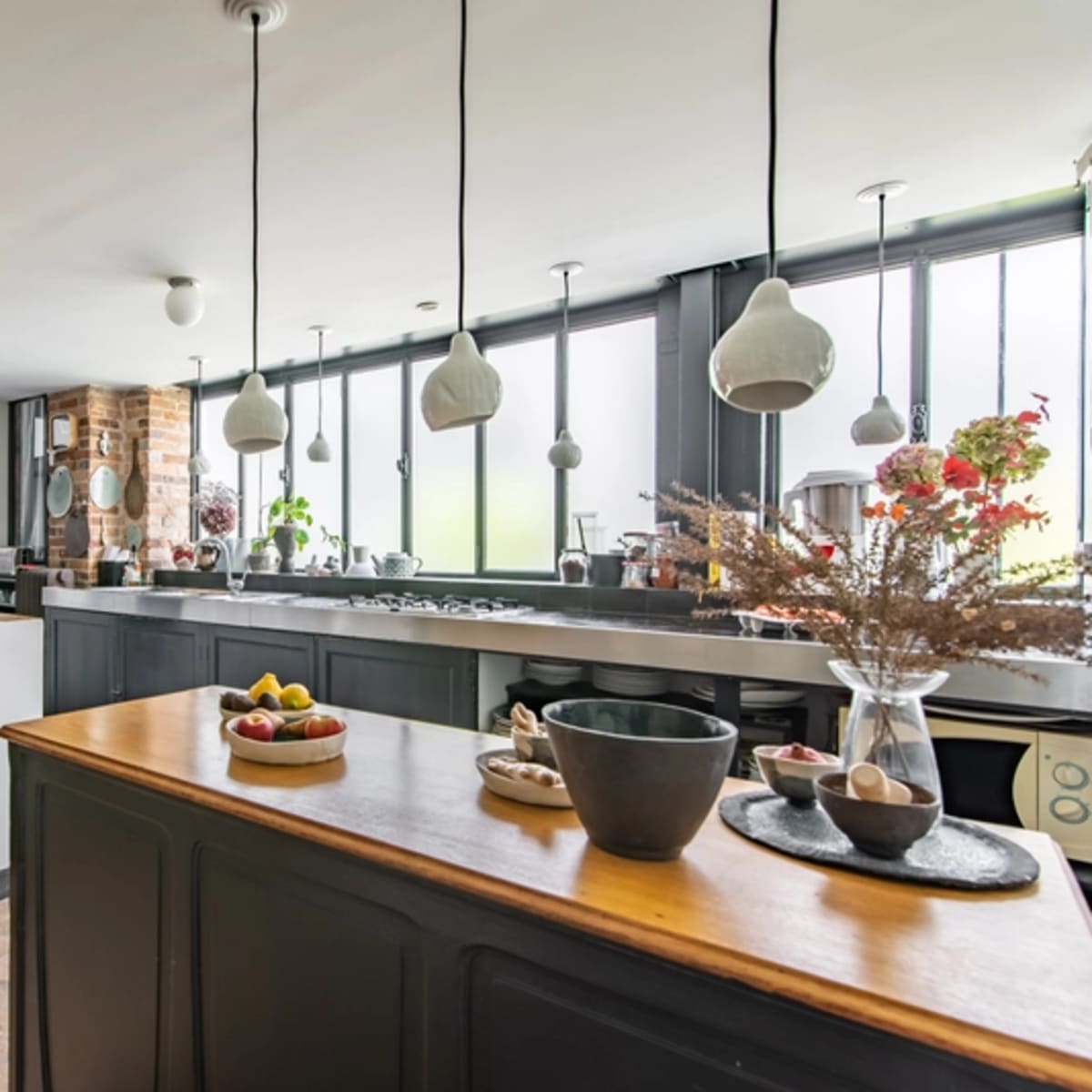 A modern kitchen features a central island with a wooden countertop, pendant lights above, and large windows providing natural light.
