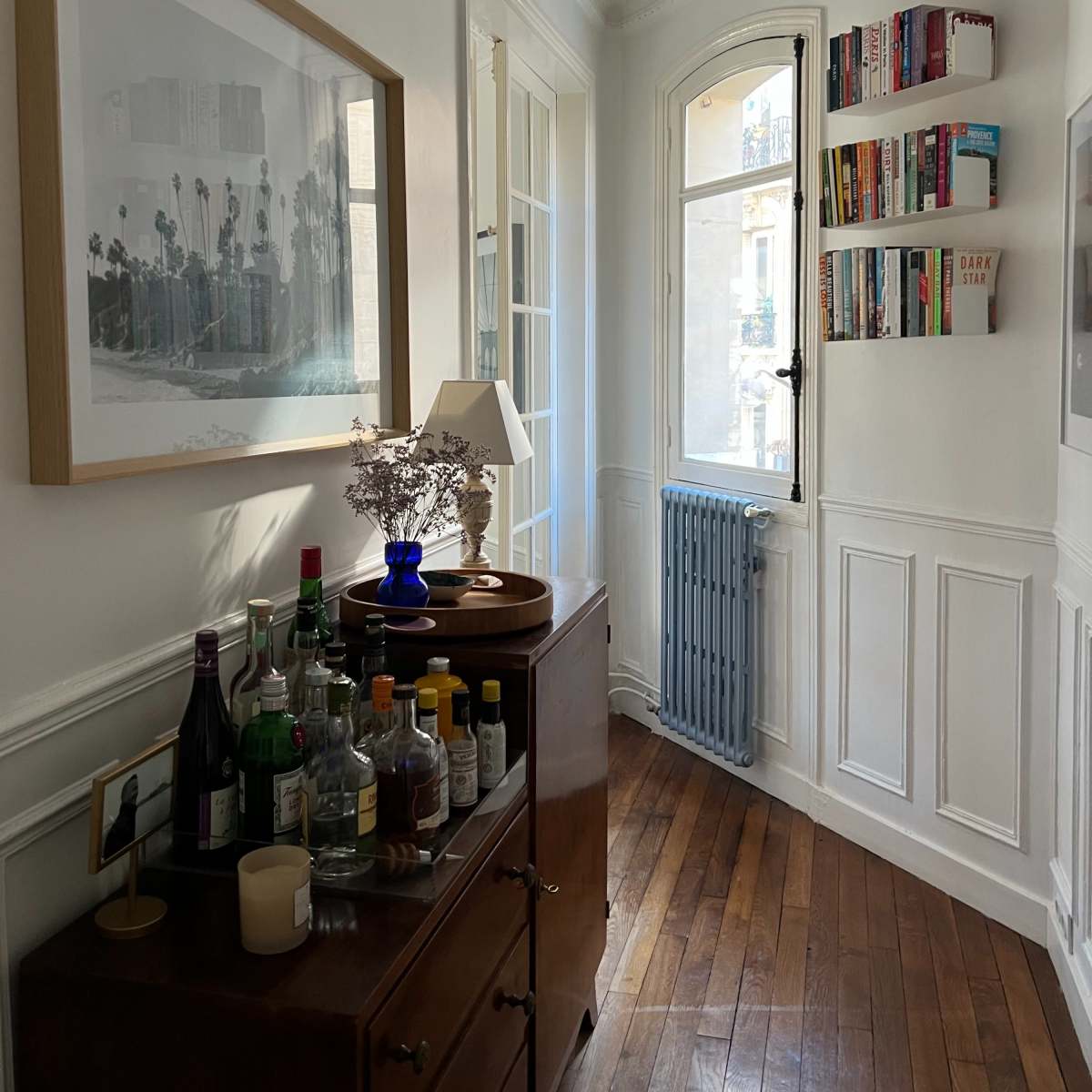 A well-lit hallway with a framed photograph on the wall, a wooden cabinet with various bottles and a lamp, and a bookshelf mounted beside a large window.