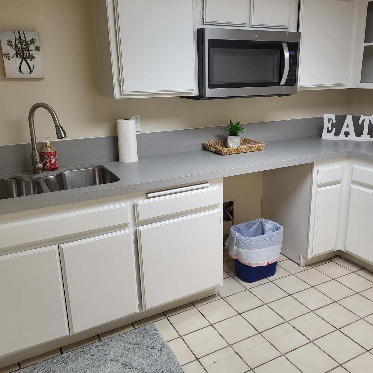 A clean kitchen featuring a sink, a microwave above the counter, and white cabinets with a decorative sign that says "EAT."