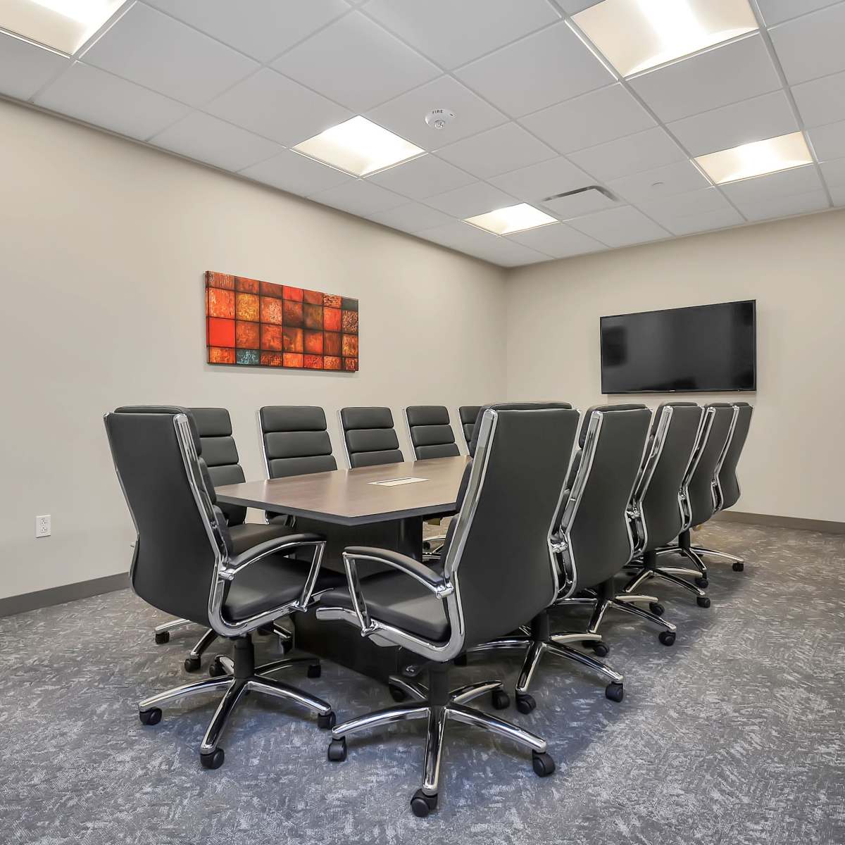 A modern conference room features a long table surrounded by black swivel chairs, with a wall-mounted screen and abstract art on the adjacent wall.