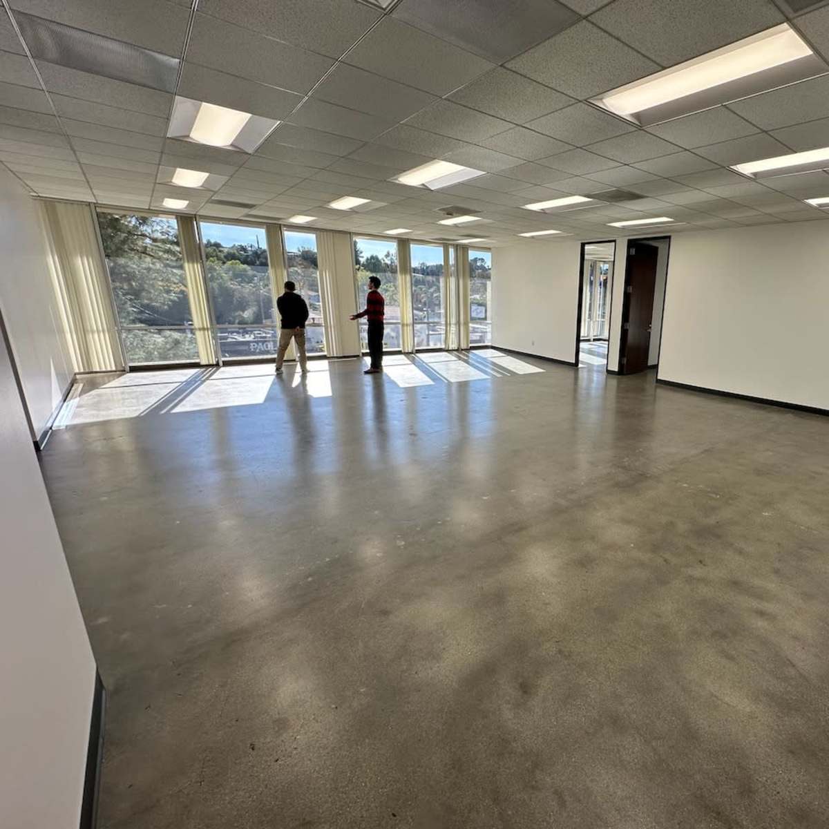 A spacious, unoccupied office room with large windows and a polished concrete floor, featuring two people standing near the windows.