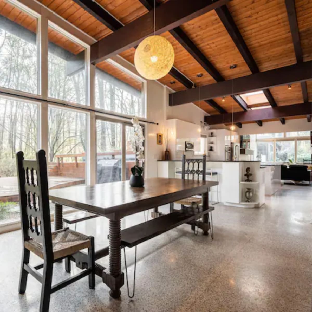 A dining area with a wooden table and chairs, large windows overlooking a wooded view, and a modern kitchen in the background.