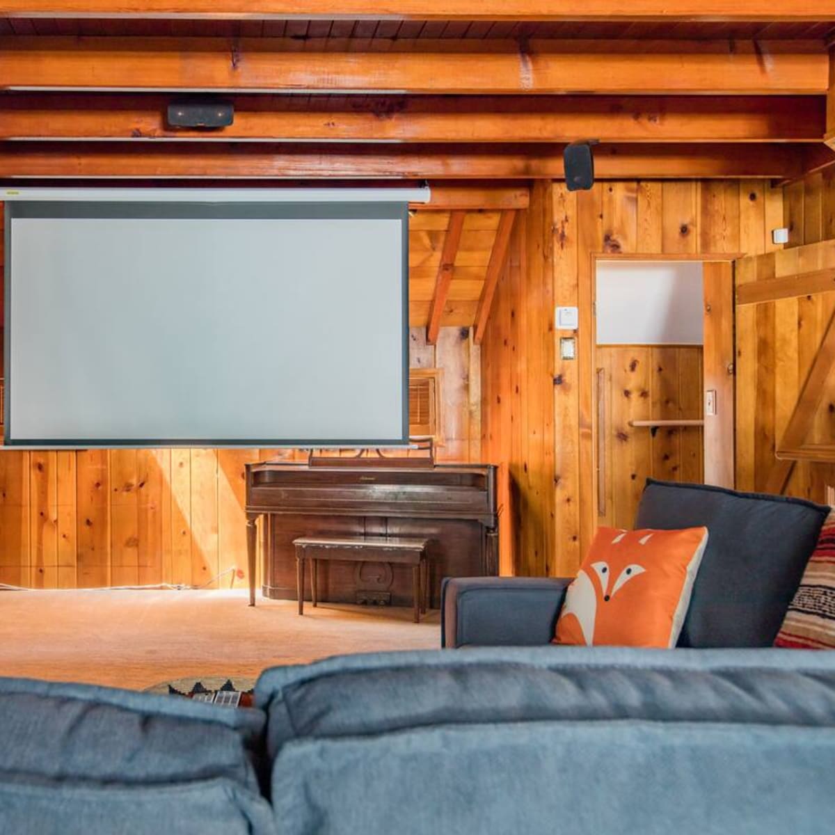 A rustic living room with wooden walls, a large projection screen, a piano, and comfortable seating.