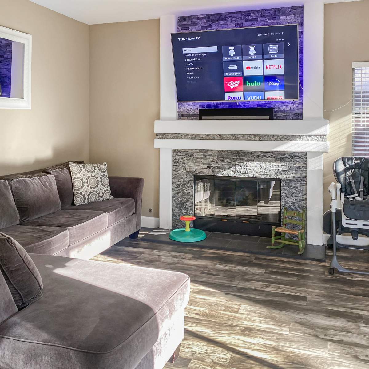 A living room with a dark sectional sofa, a stone fireplace, a television mounted on the wall above, and a high chair near a window.