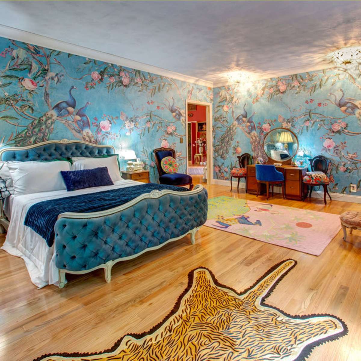 A brightly decorated bedroom featuring a blue floral wall mural, a tufted bed, an ornate dresser, and a tiger-striped rug on the wooden floor.