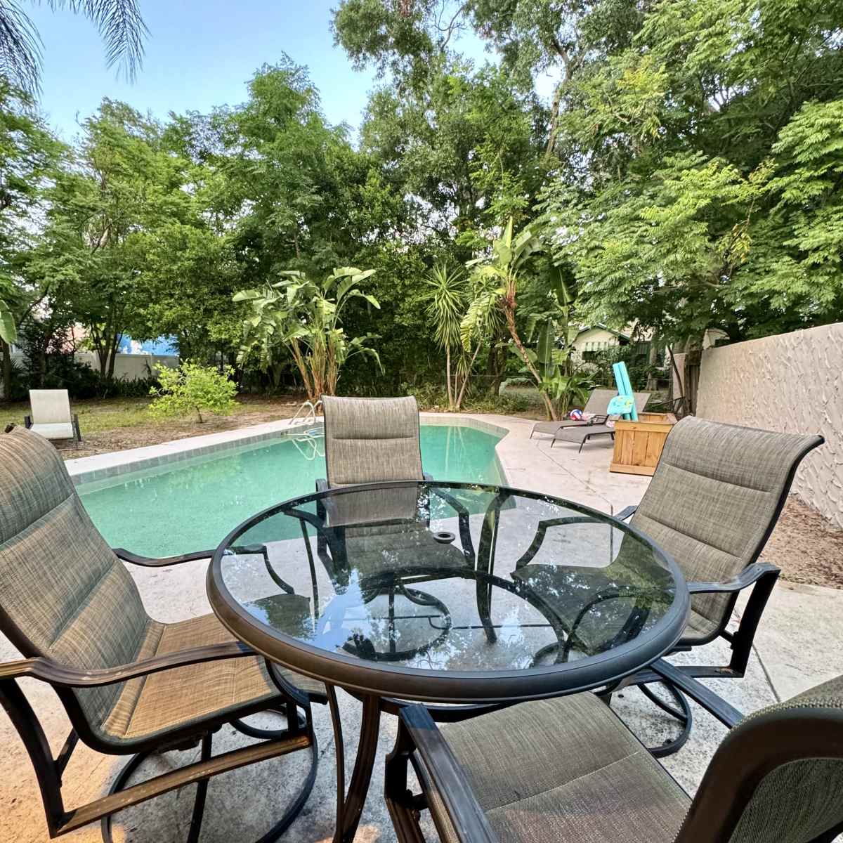 A round glass table with four chairs sits on a patio overlooking a swimming pool surrounded by greenery.