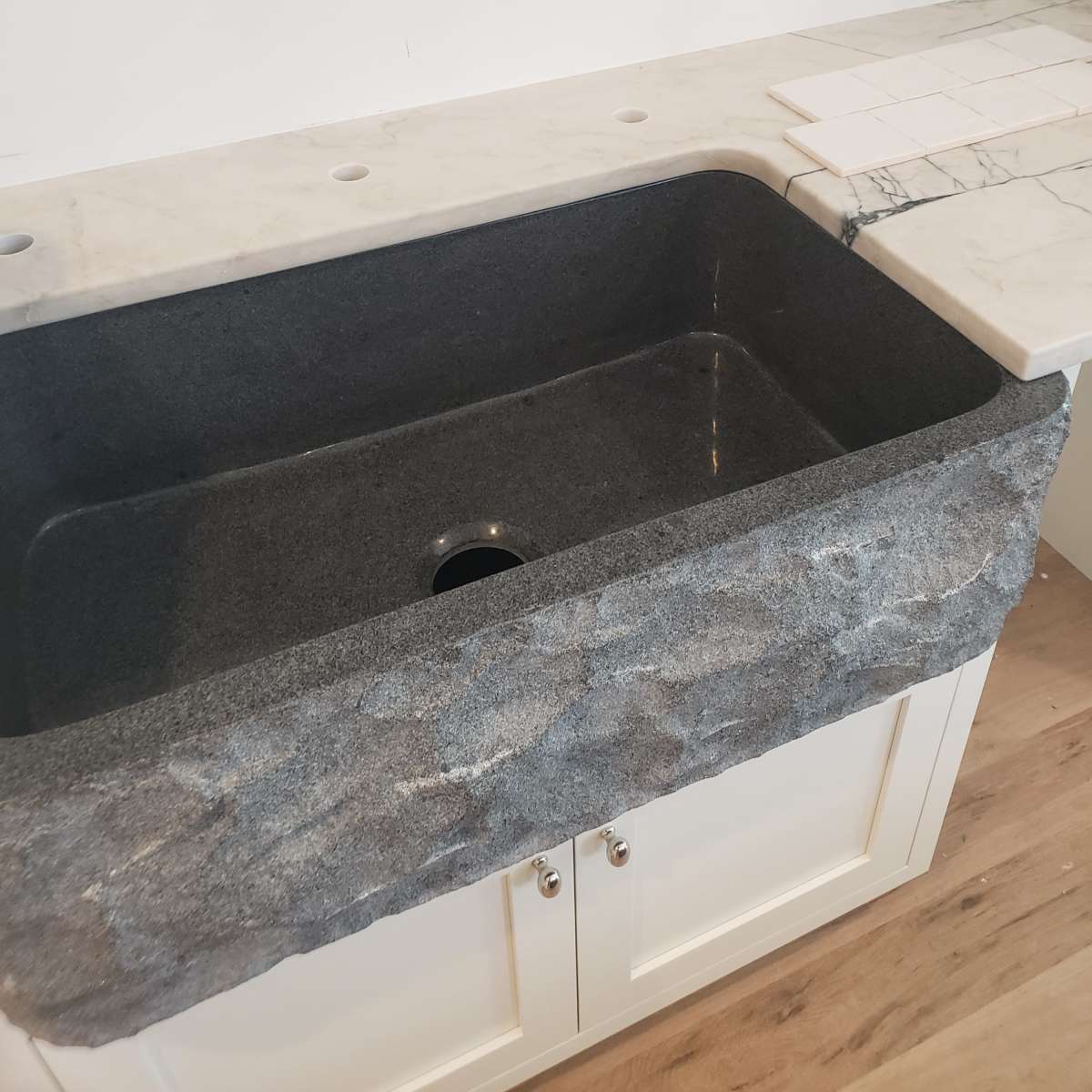 A dark granite farmhouse sink integrated into a light-colored countertop with a backsplash and unfinished wall electrical outlets visible.