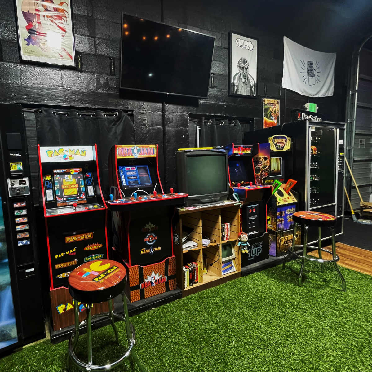 An arcade corner featuring vintage gaming machines, a television, and a vending machine, all set against a black wall with artificial turf.