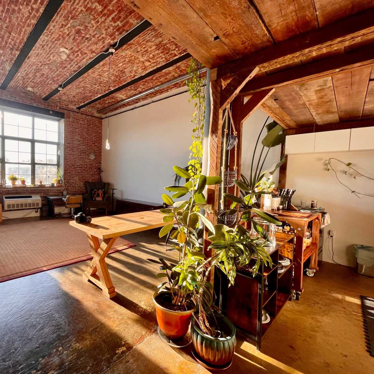 An industrial-style loft with exposed brick walls, wooden beams, and a large window allowing natural light to fill the space, featuring a seating area, a dining table, and potted plants.