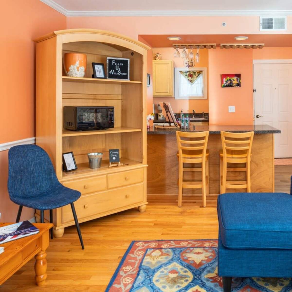 A cozy living space with an orange wall, a blue sectional sofa, a wooden shelf unit, and a kitchen area in the background.