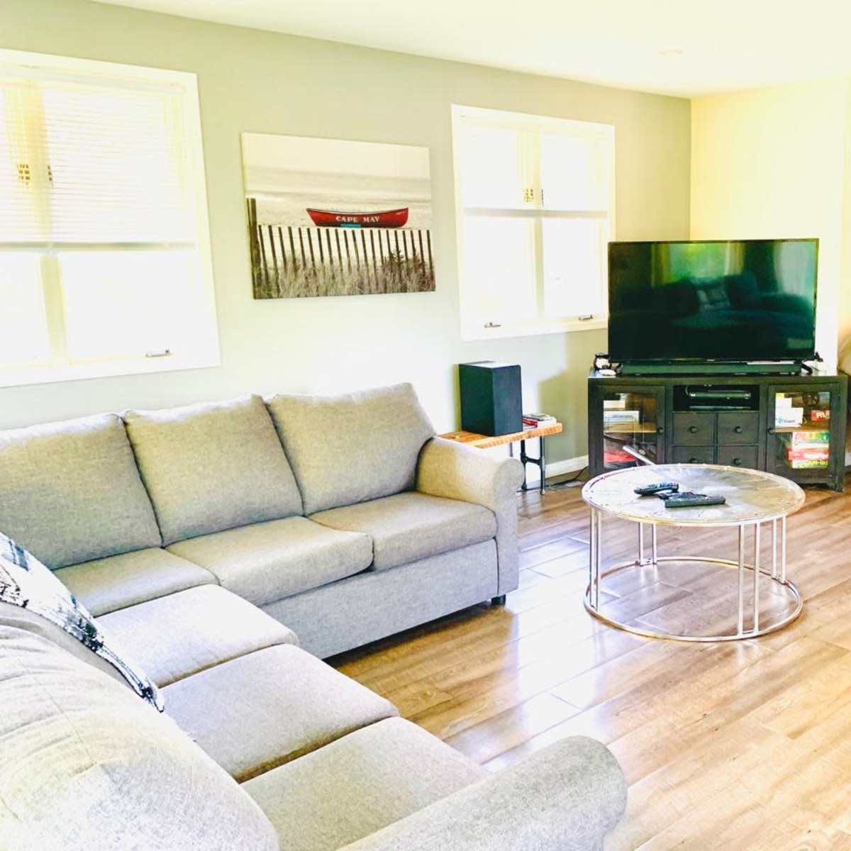 A living room features a gray sectional sofa, a glass coffee table, and a TV on a stand, with light streaming in from windows.