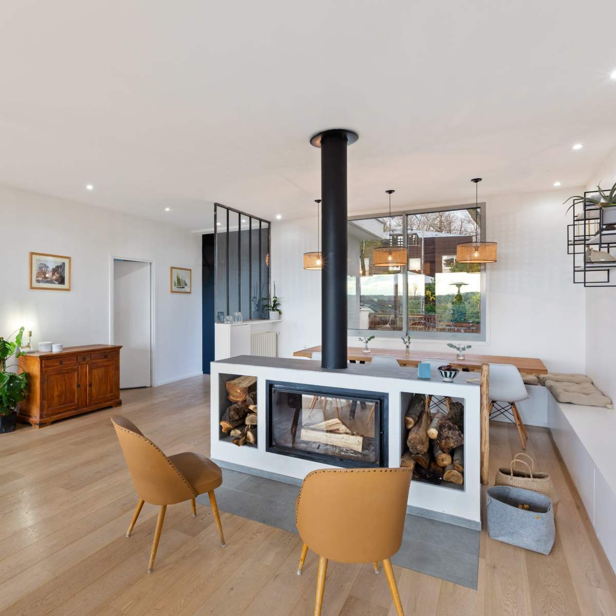A contemporary living space featuring a centrally located fireplace, a dining area with a long table, and a staircase leading to an upper level.