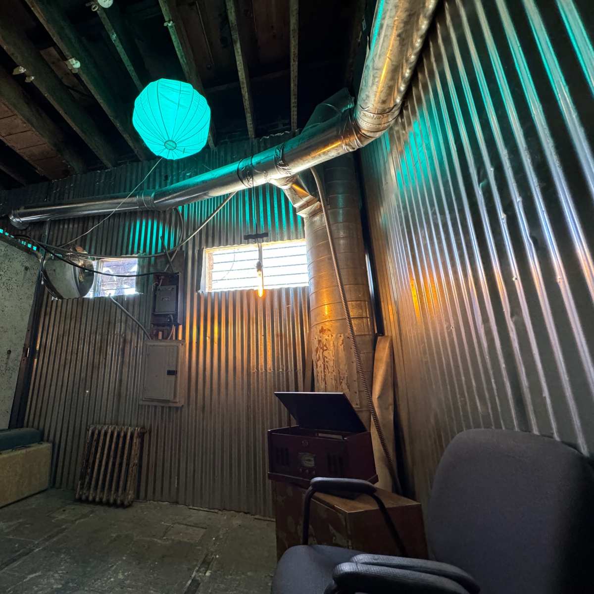 A room with corrugated metal walls, a teal lantern hanging from the ceiling, and a vintage record player near a chair.
