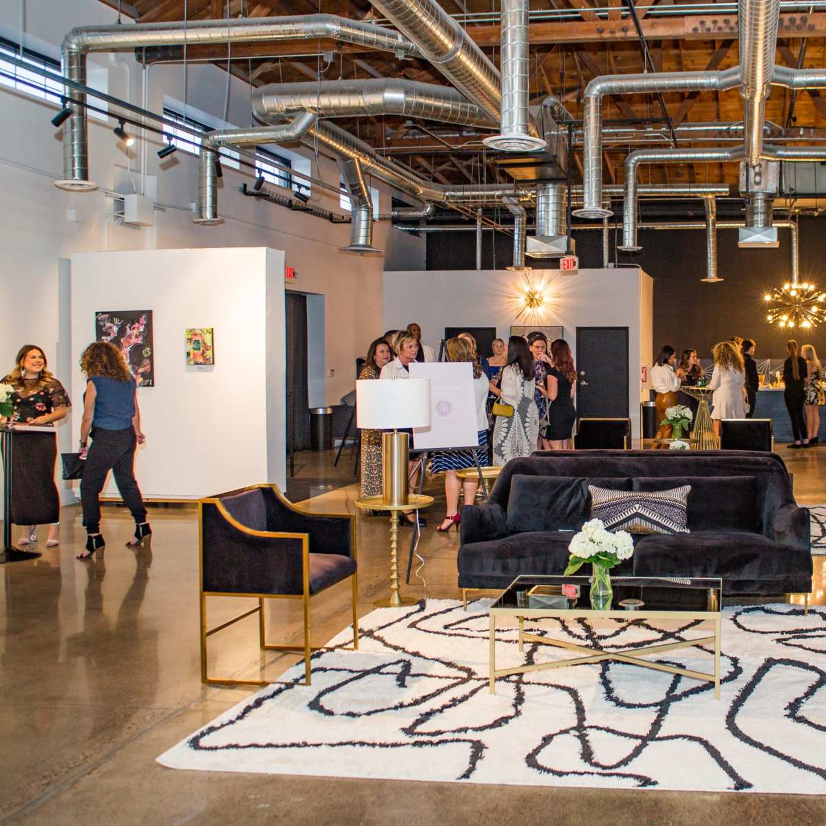 A modern gallery space with a stylish black seating area, a patterned rug, and several people attending an event, with artwork displayed on the walls.