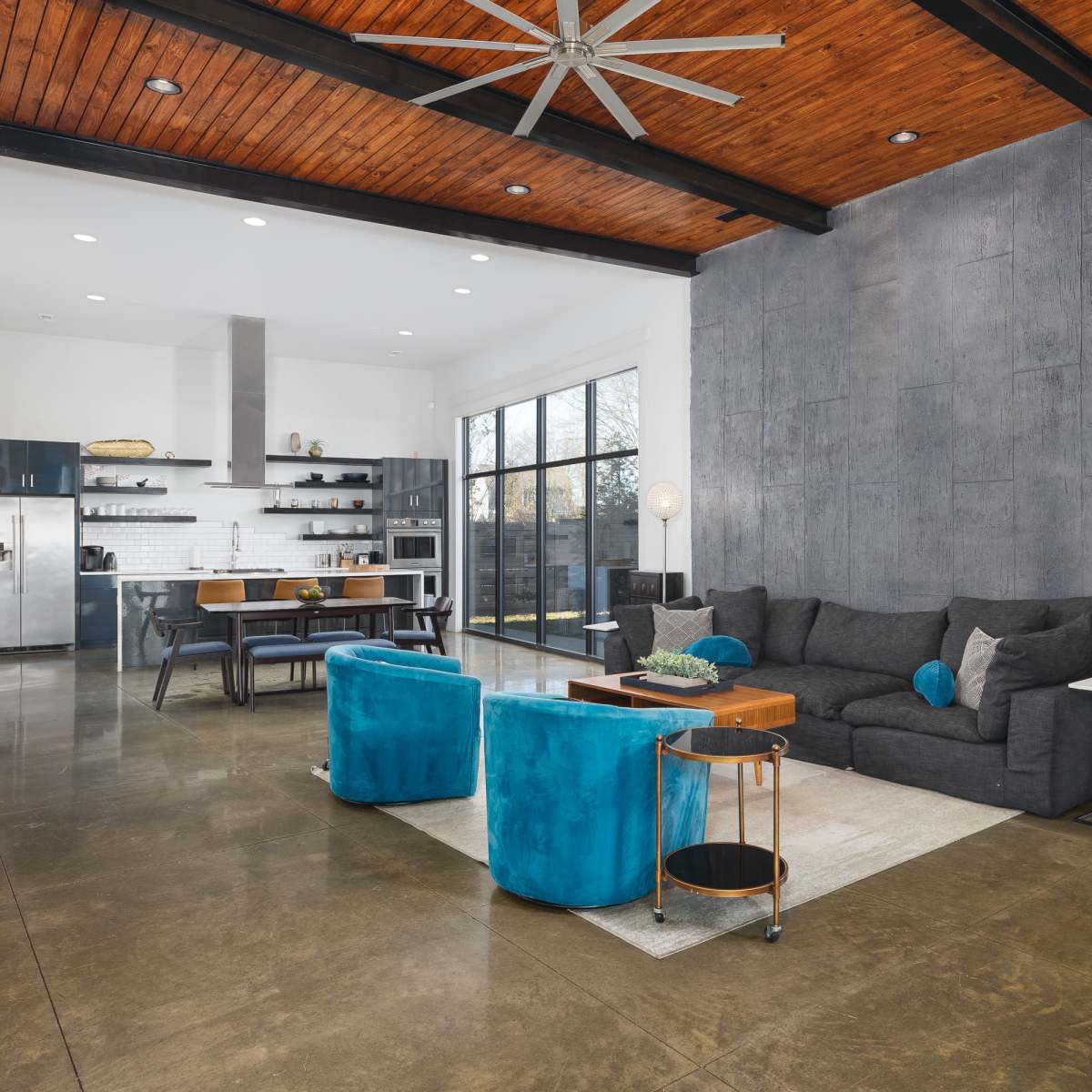 A modern living room featuring a large dark sectional sofa, two vibrant blue chairs, a wooden table, and a kitchen area with sleek appliances.