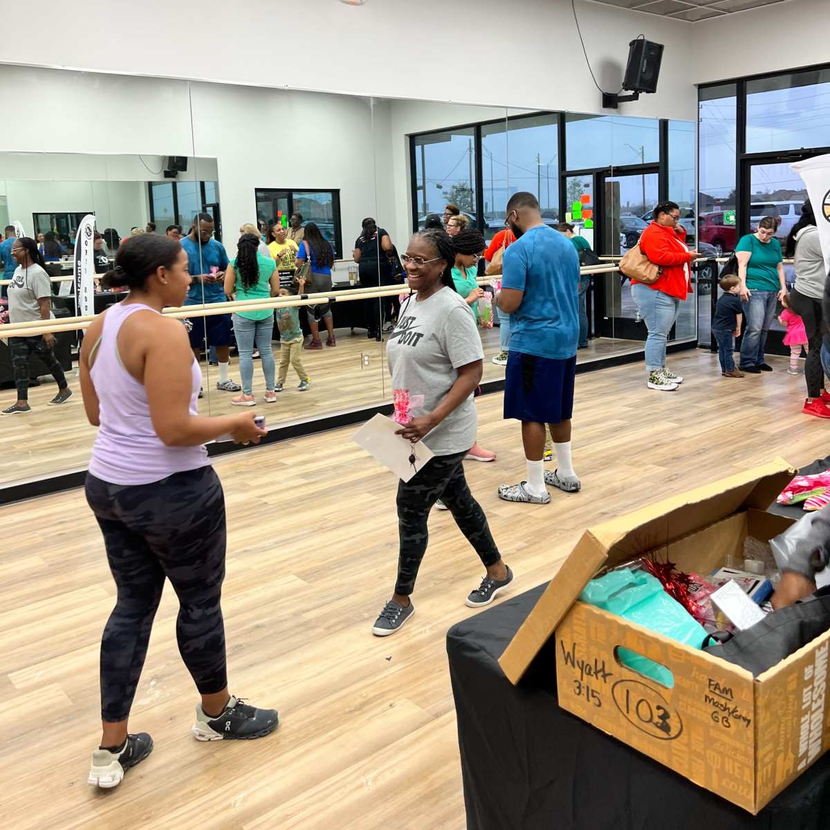 A group of people gather in a well-lit fitness studio with mirrors, some participating in an exercise class and others waiting around, while a box of supplies sits on a table.