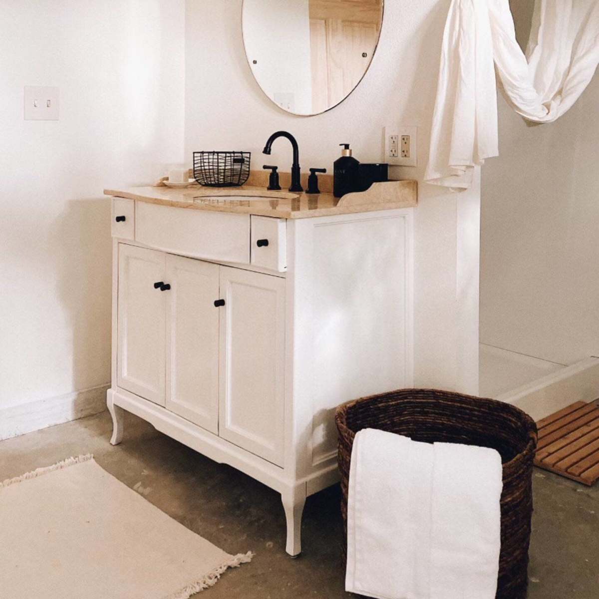 A bathroom featuring a white vanity with a round mirror, a black faucet, and a woven basket alongside a hanging shower curtain.