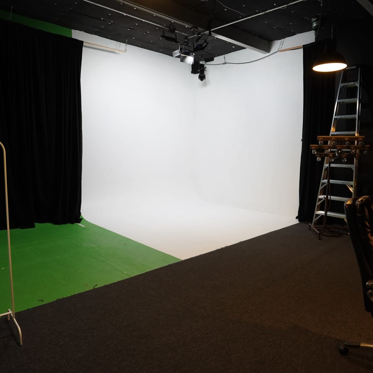 A photography studio with a white backdrop, a black curtain, a green section of the floor, and a black office chair nearby.