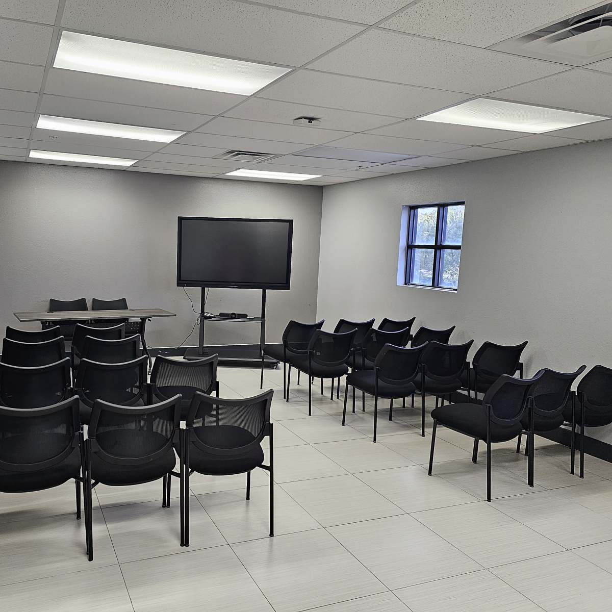 An empty meeting room with rows of black chairs facing a table and a wall-mounted screen.