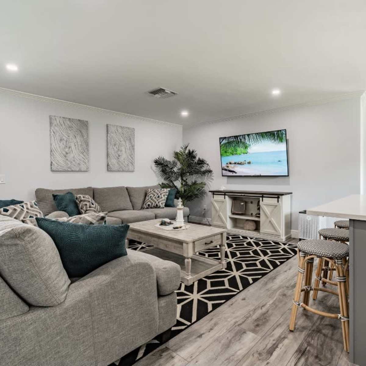 A modern living room features a large gray sectional sofa, a coffee table, and a flat-screen TV displaying a scenic beach image, with a kitchen area visible in the background.