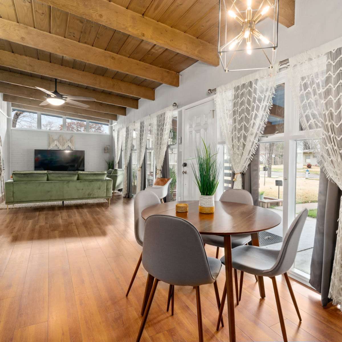 A dining area features a round table surrounded by chairs, with large windows showcasing a living space adorned with a green sofa and a television.