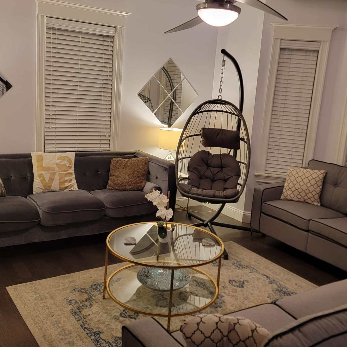 A cozy living room features a hanging chair, two gray sofas, a round glass coffee table, and decorative accents, with natural light filtering through the windows.