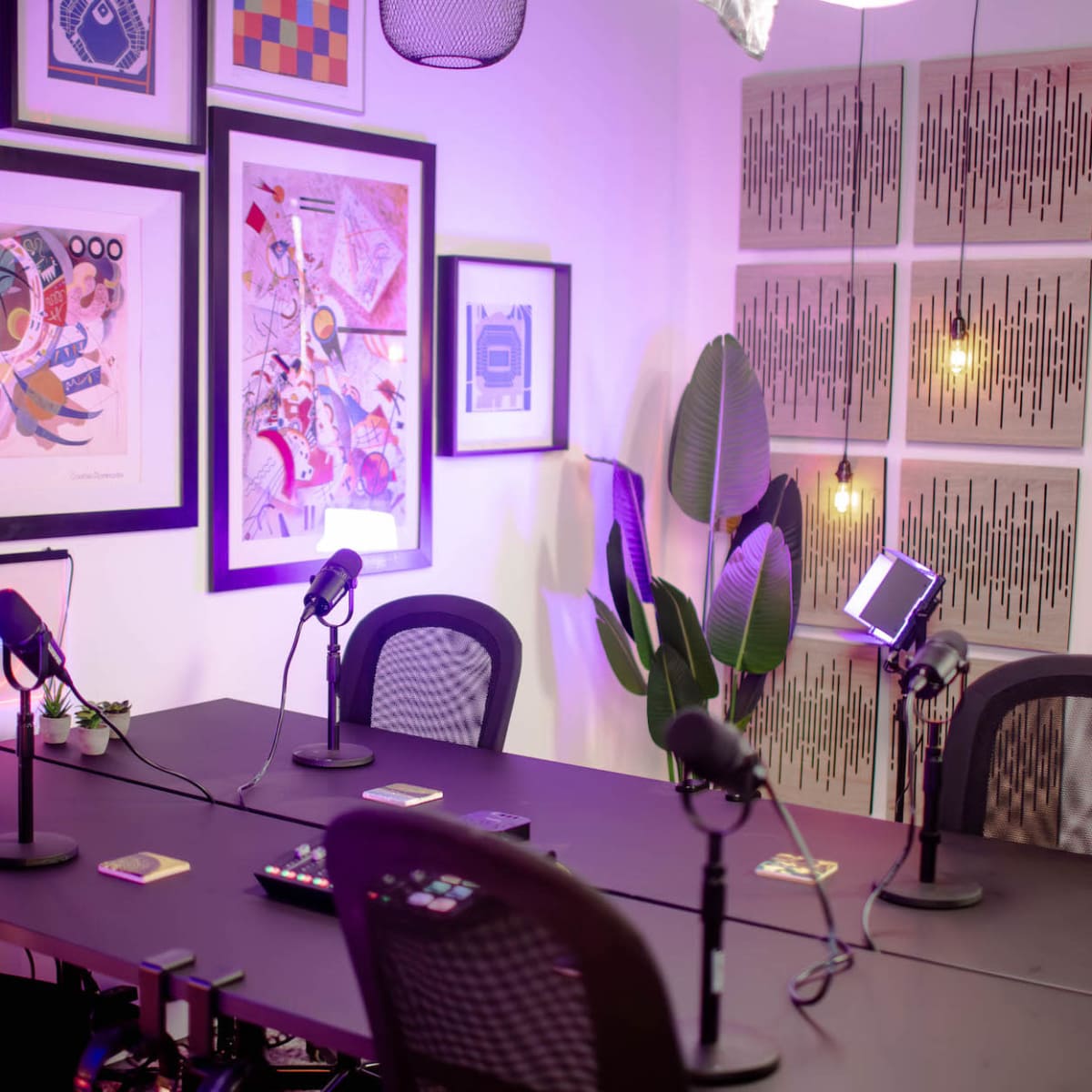 A modern podcast studio with a table, four microphones, framed artwork on the walls, and a decorative plant in the corner.