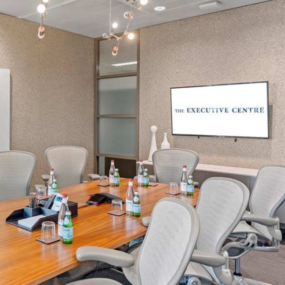 A modern conference room with a large wooden table, eight ergonomic chairs, a whiteboard, and a wall-mounted monitor displaying "The Executive Centre."