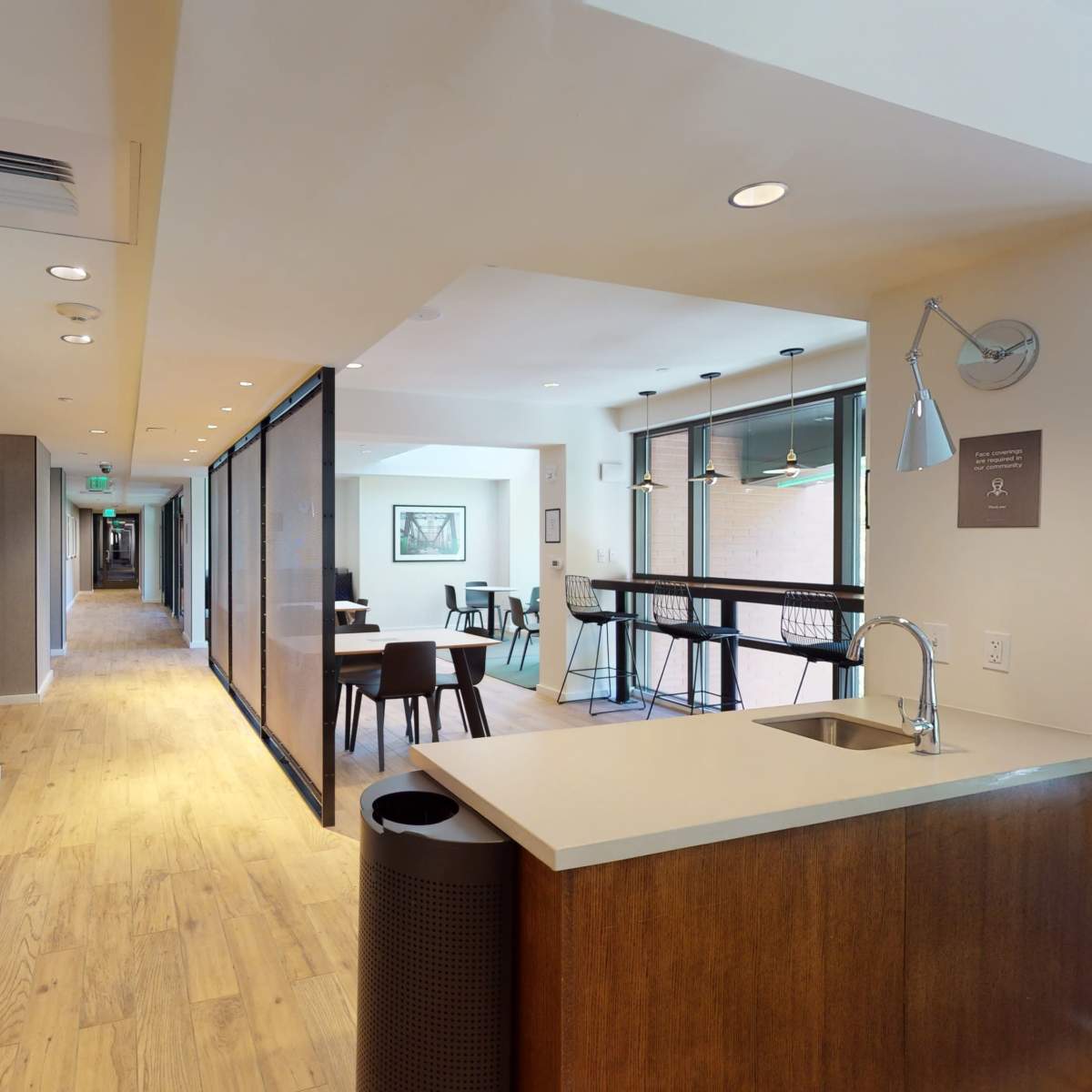A modern hallway that connects a kitchen area to a seating area, featuring a mix of wooden flooring and light-colored walls, with glass panel dividers.