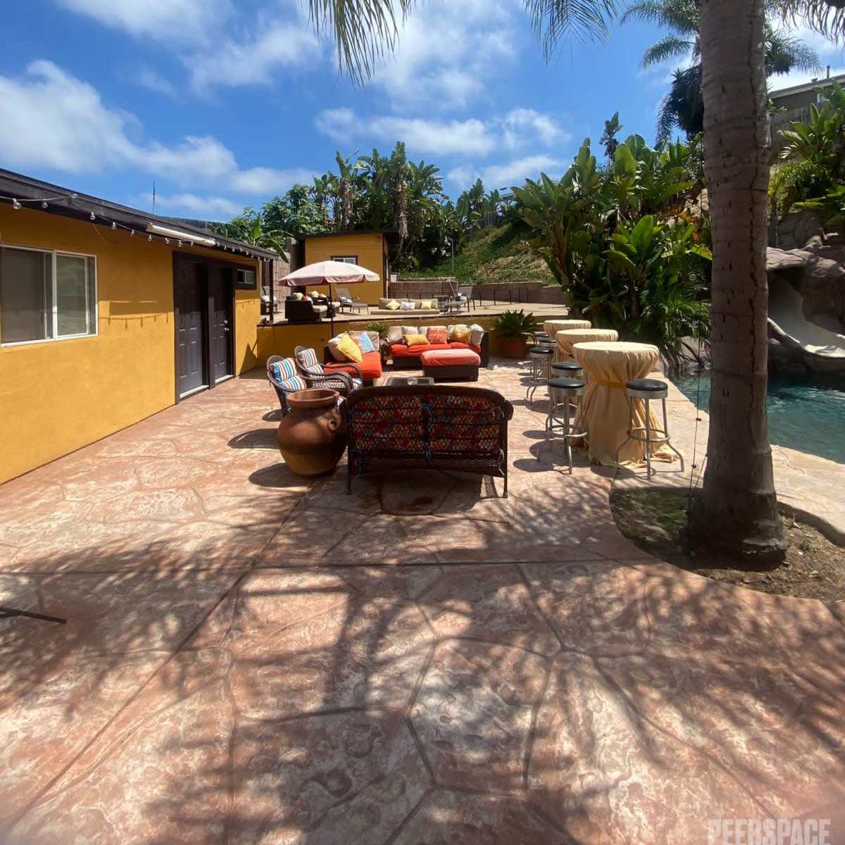 A patio area with colorful seating and a pool surrounded by tropical plants under a partly cloudy sky.