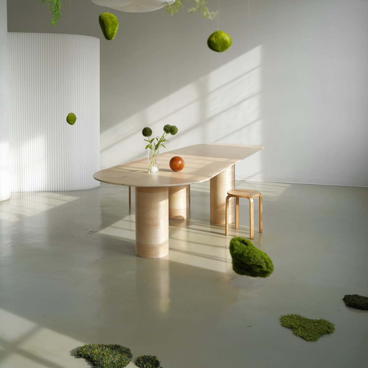 A minimalist interior features a large wooden table with a single stool, surrounded by suspended moss and a few decorative elements on the floor.