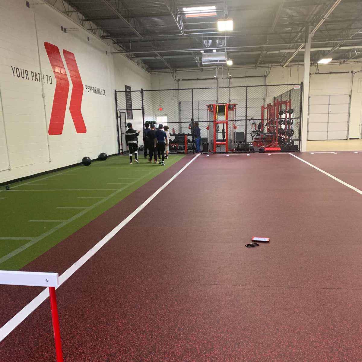 An indoor training facility with a red and black rubber floor, turf, exercise equipment, and a group of athletes lined up near the wall.