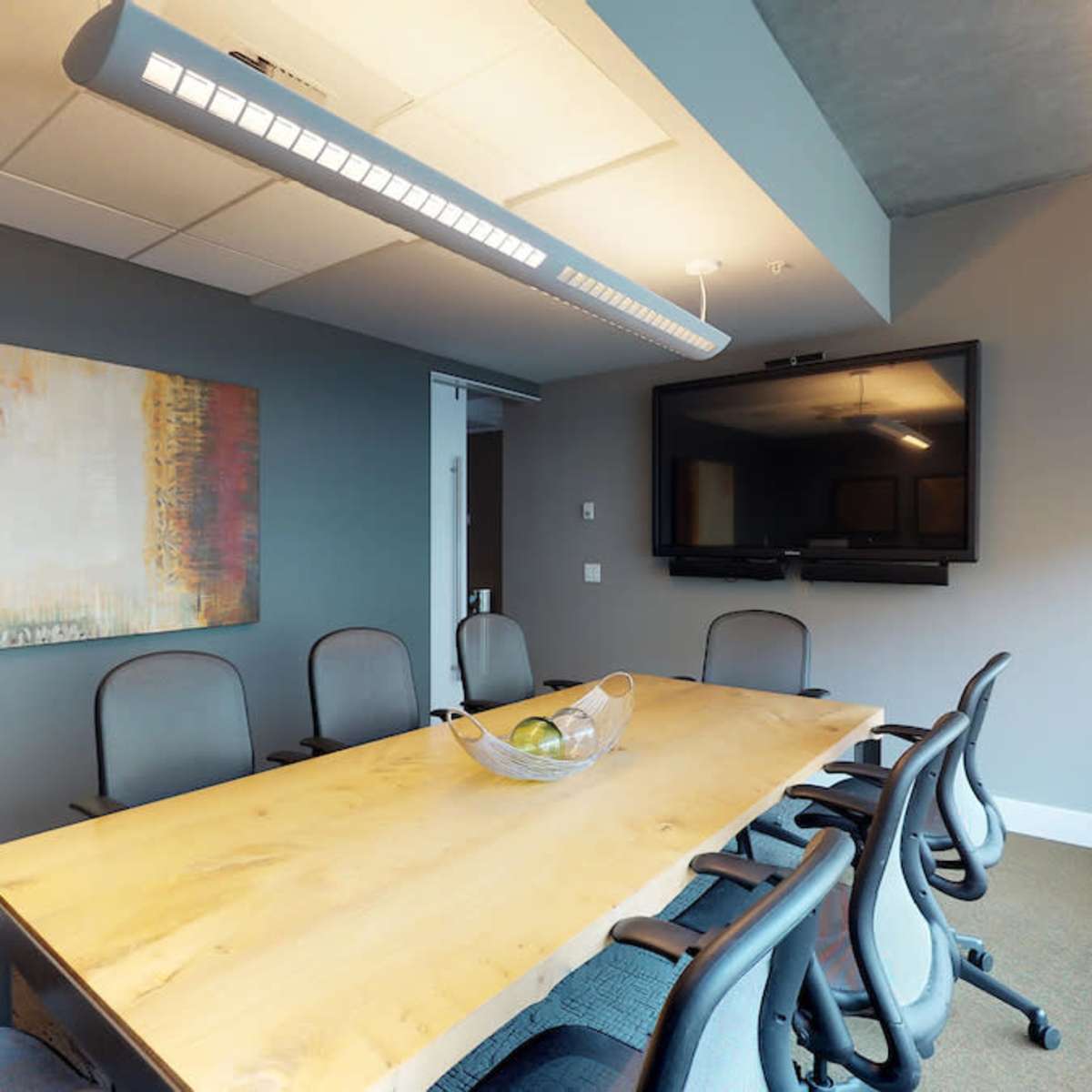 A modern conference room features a large wooden table surrounded by ergonomic chairs, with a wall-mounted TV and a piece of abstract artwork.