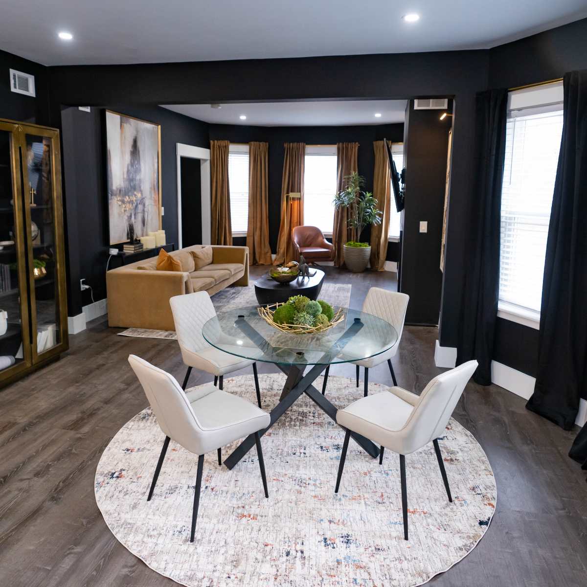 A modern living space features a glass dining table with four chairs, surrounded by a cozy seating area and large windows dressed in dark curtains.