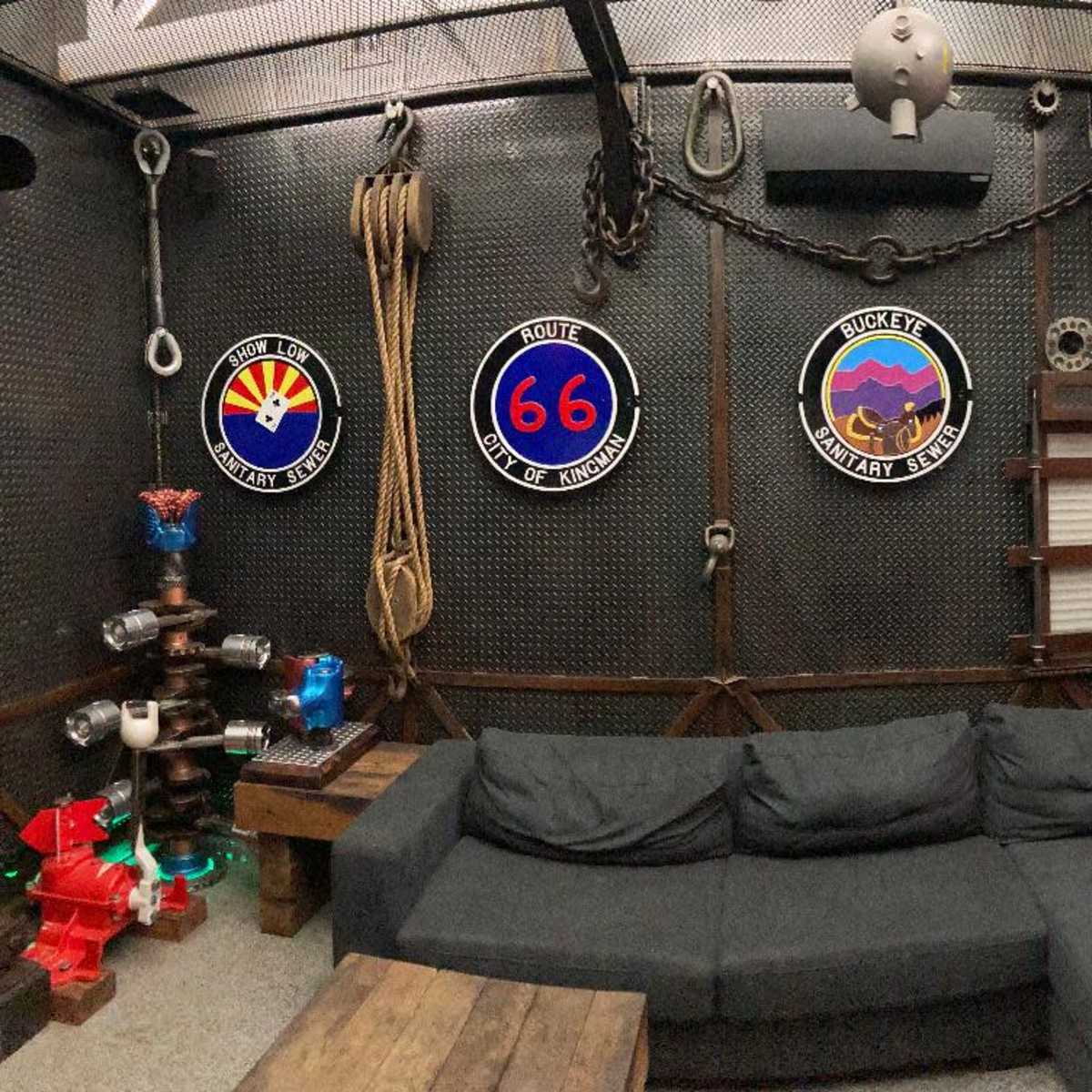 A room with a dark, industrial design featuring a seating area with a large sofa, adorned walls with military-themed plaques, and various ropes and chains hanging from the ceiling.