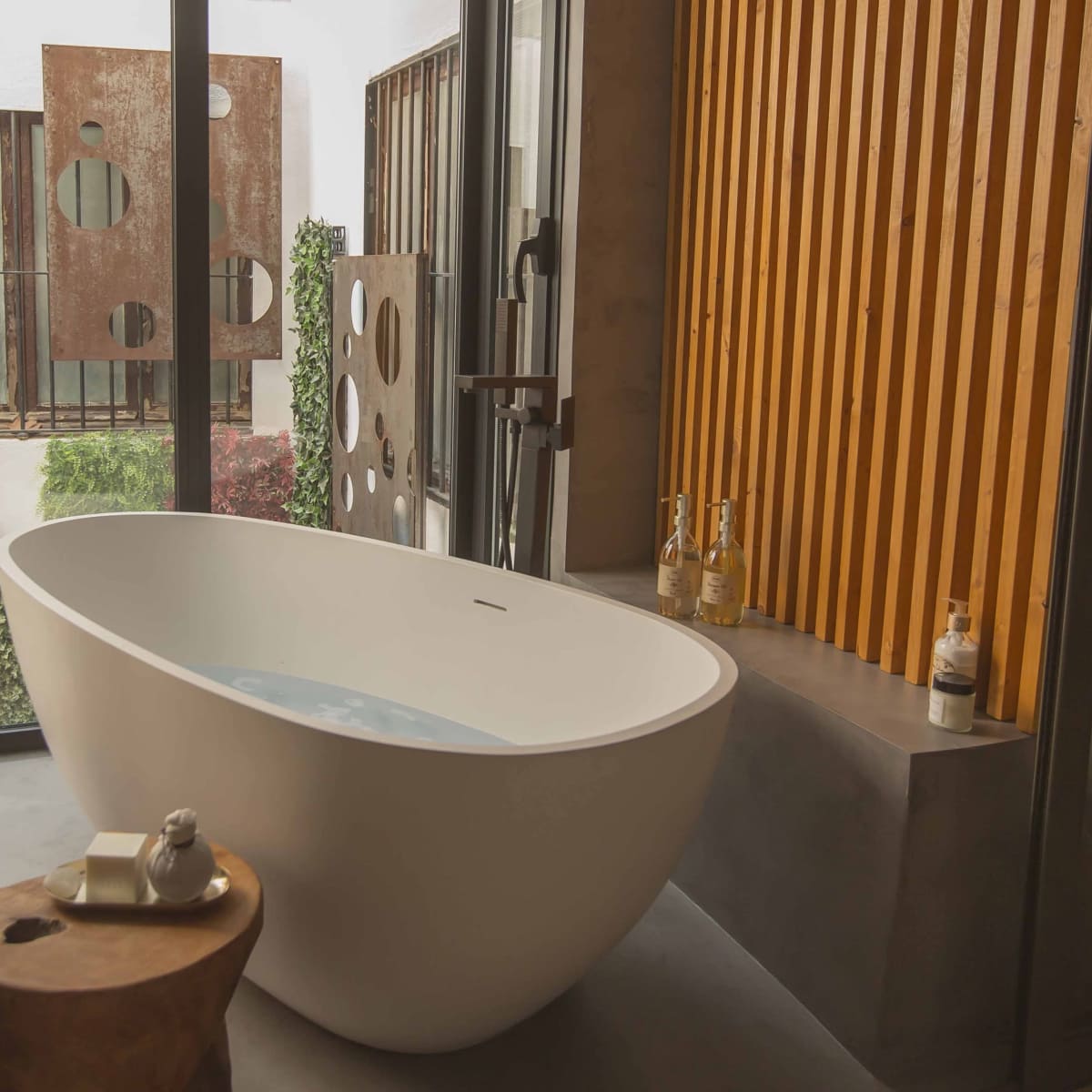 A modern bathroom featuring a freestanding white bathtub beside a brown wooden stool, with large windows providing a view of outdoor greenery.