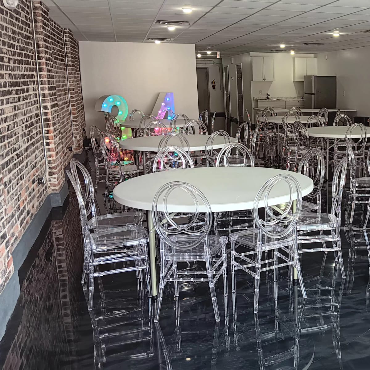 A spacious, modern event room featuring several round tables with transparent chairs and a large kitchen area in the background.