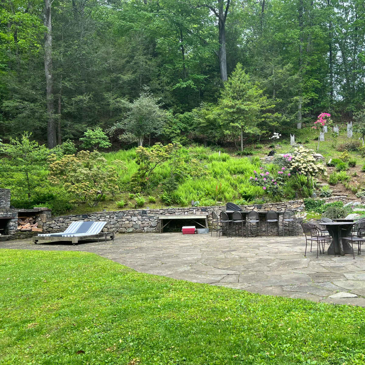 A stone patio with a circular table and chairs is surrounded by greenery and a hillside of flowers and shrubs.