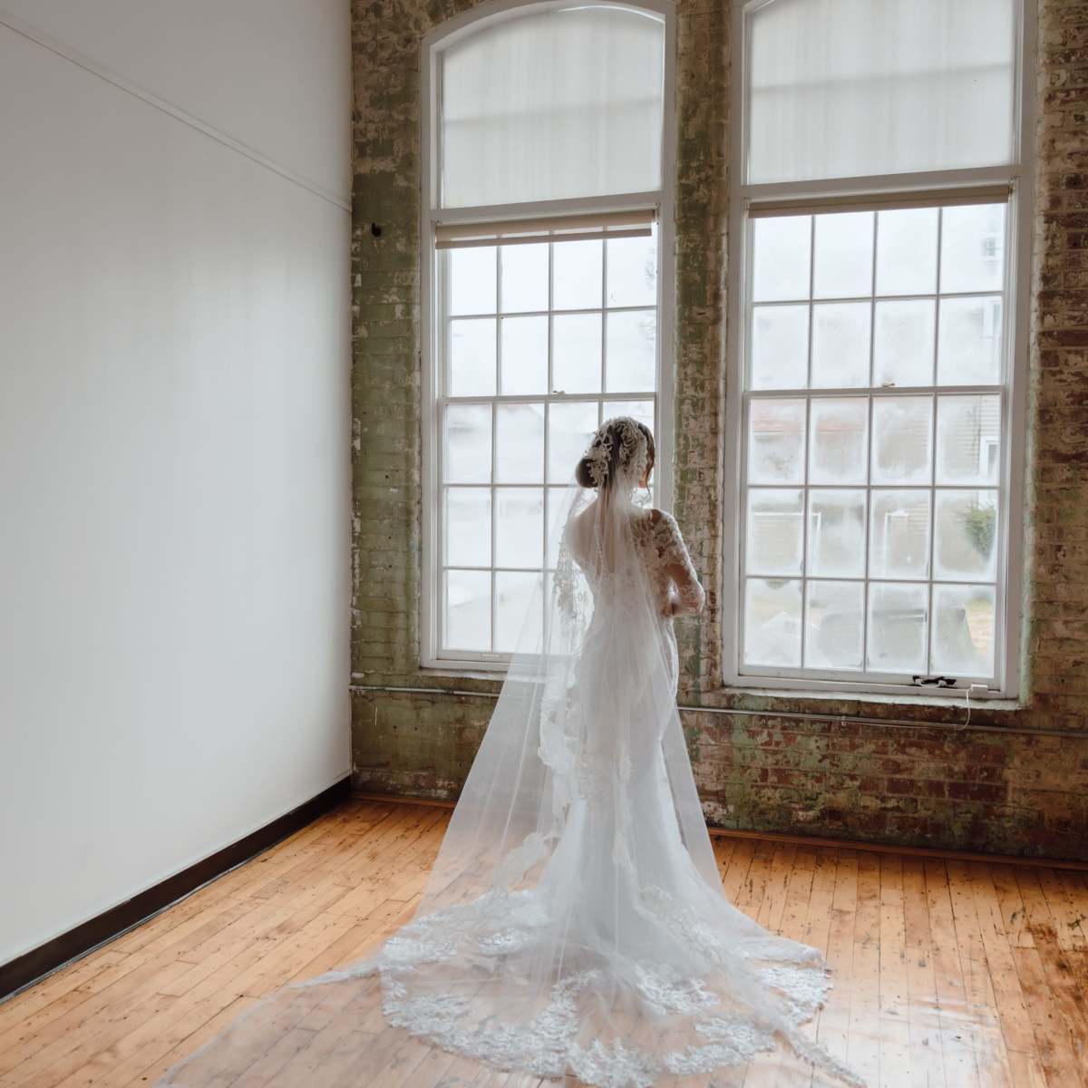 A bride in a white gown and veil stands near large windows in a room with exposed brick walls and wooden floors.
