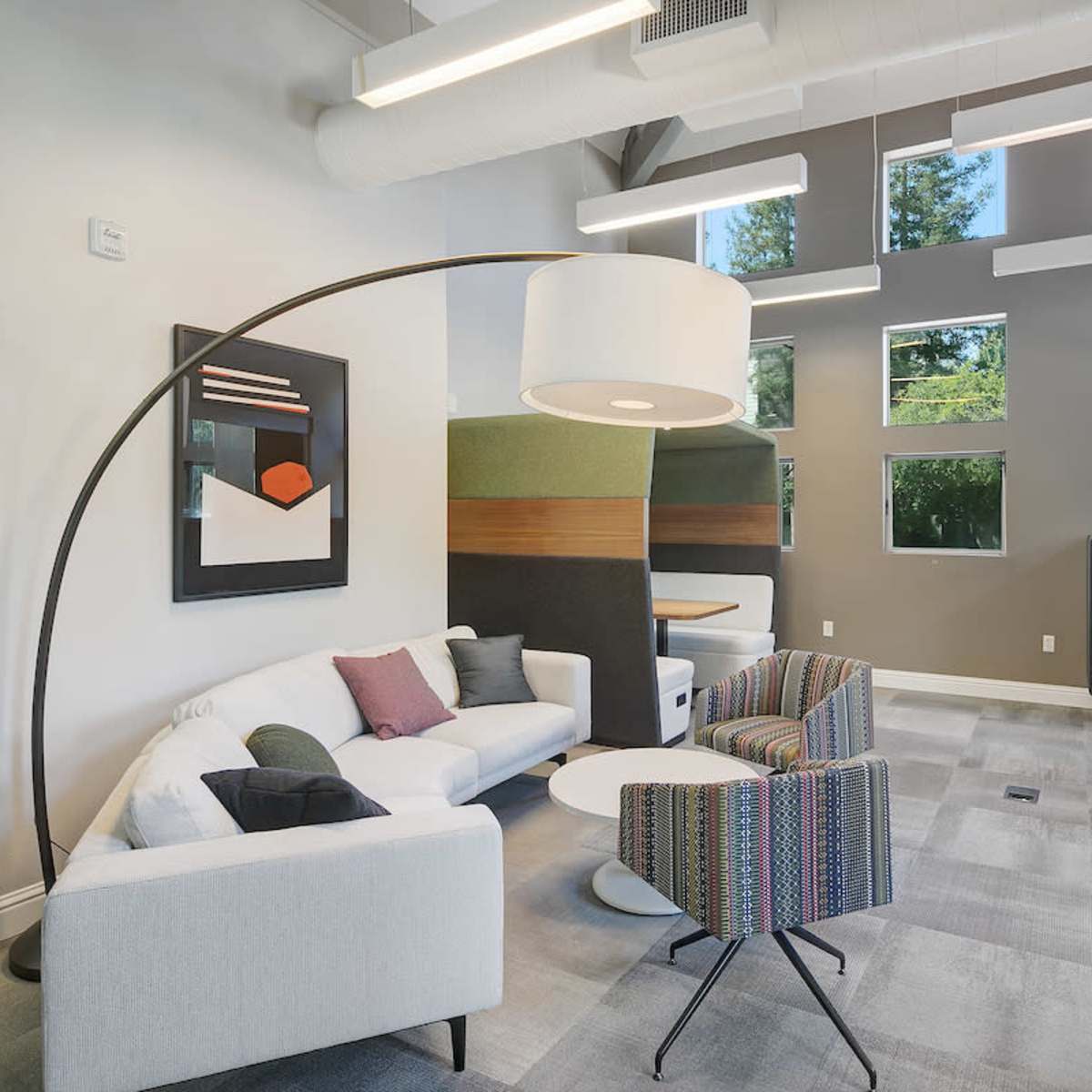A modern seating area with a curved lamp, a white couch, a colorful patterned chair, a round table, and large windows providing natural light.