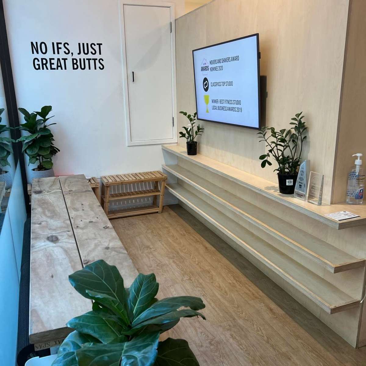 A modern waiting area with a wooden bench, potted plants, and a television mounted on the wall displaying a message.