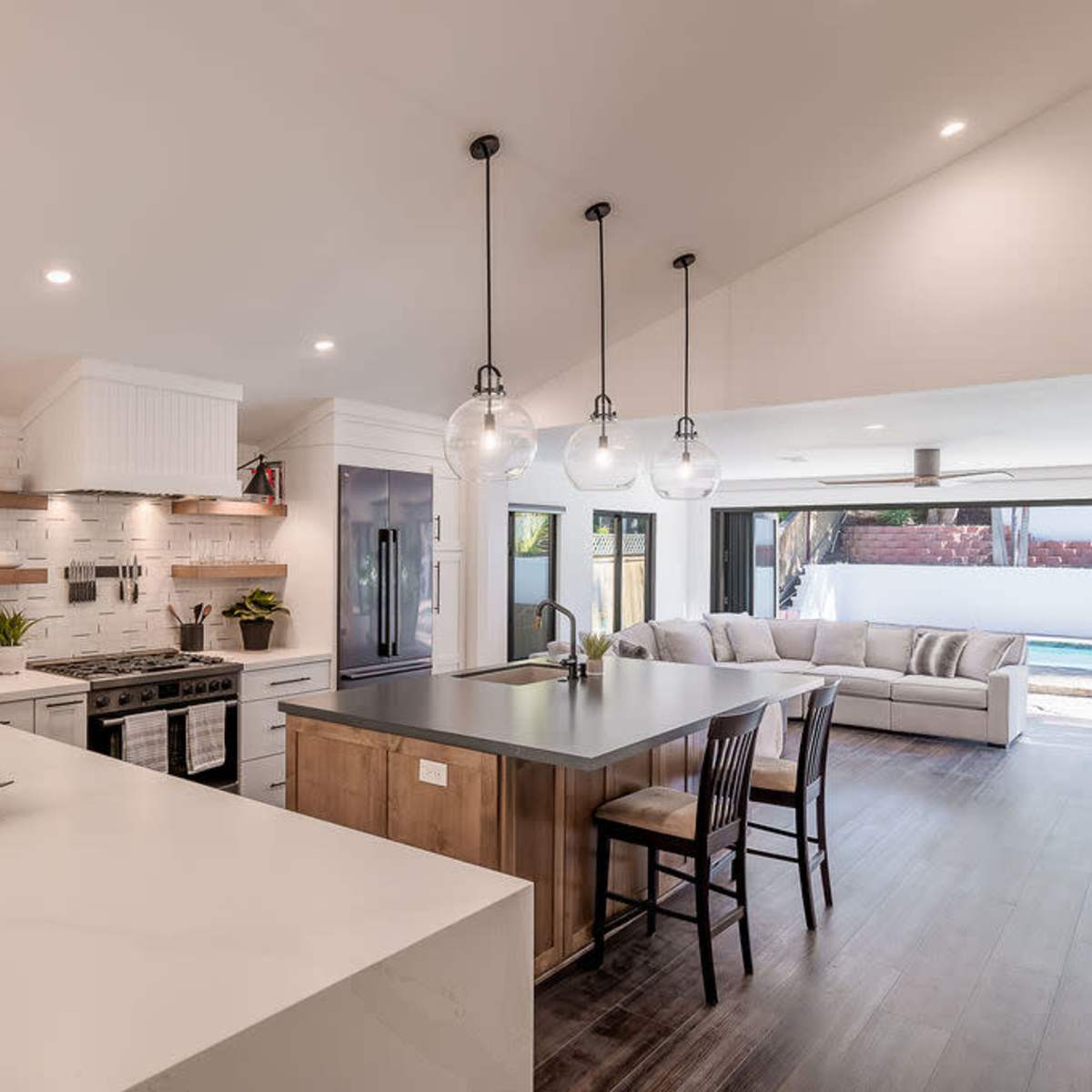 A modern kitchen features a large island, stainless steel appliances, and an open layout that connects to a living area with a sofa and a view of a pool.