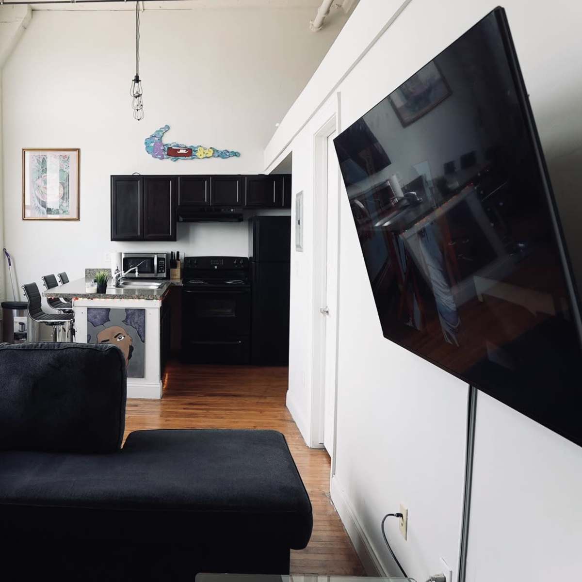 A modern living space featuring a black couch, a kitchen with dark cabinetry, and large windows allowing natural light.