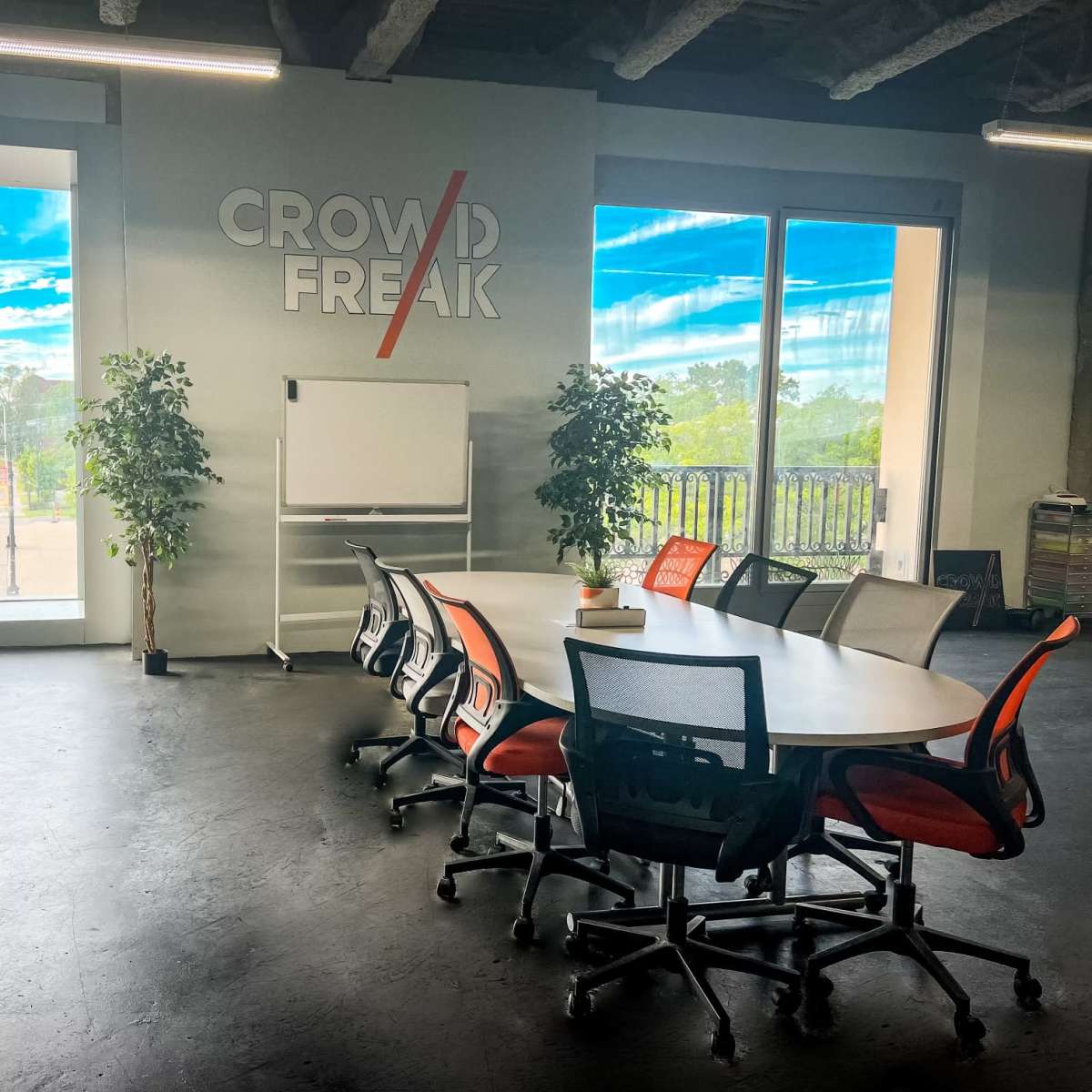 A conference room features a large round table surrounded by chairs, with floor-to-ceiling windows showing an outdoor view and a wall displaying the text "CROWD FREAK."
