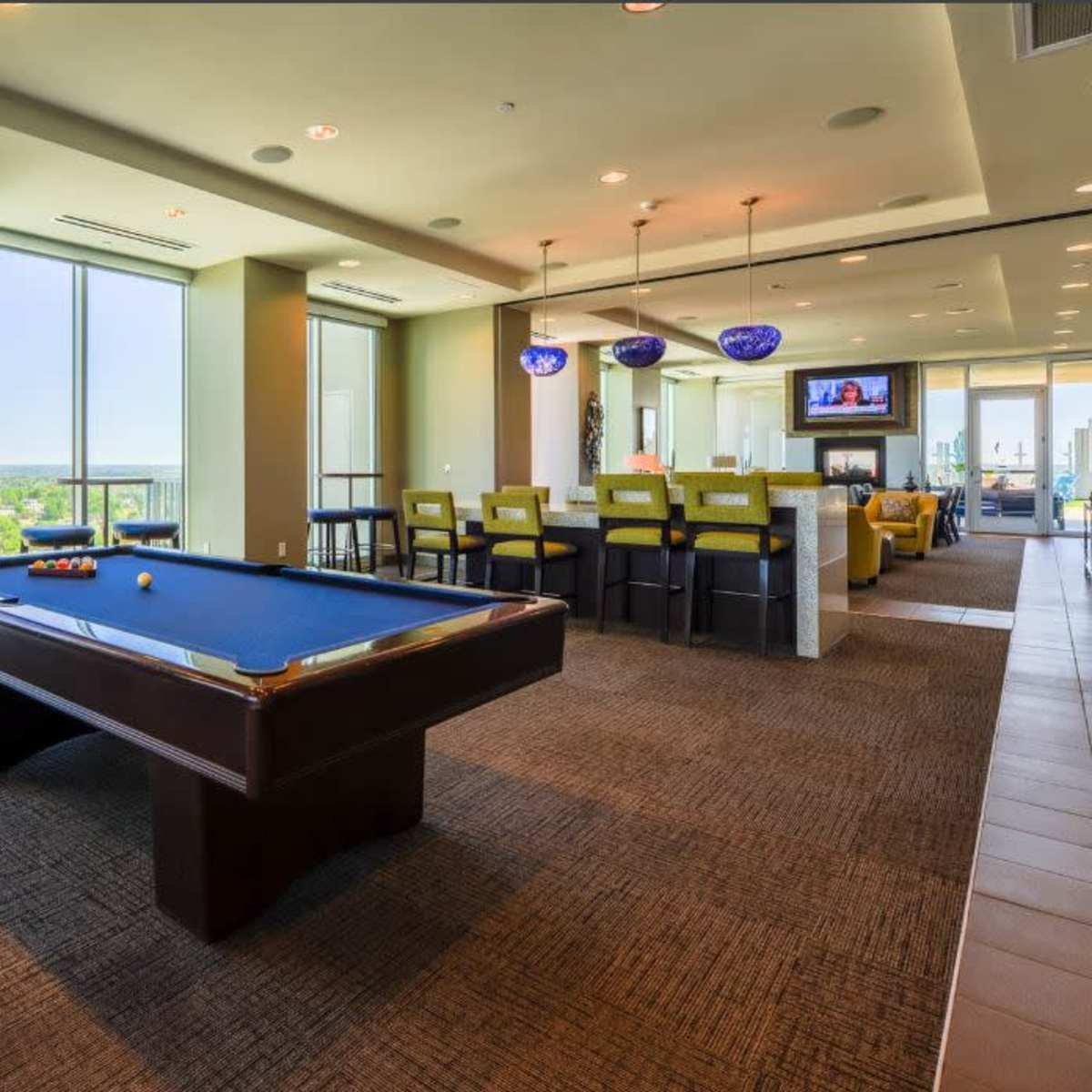 A billiards table is positioned in a well-lit lounge area with large windows showcasing a city view.